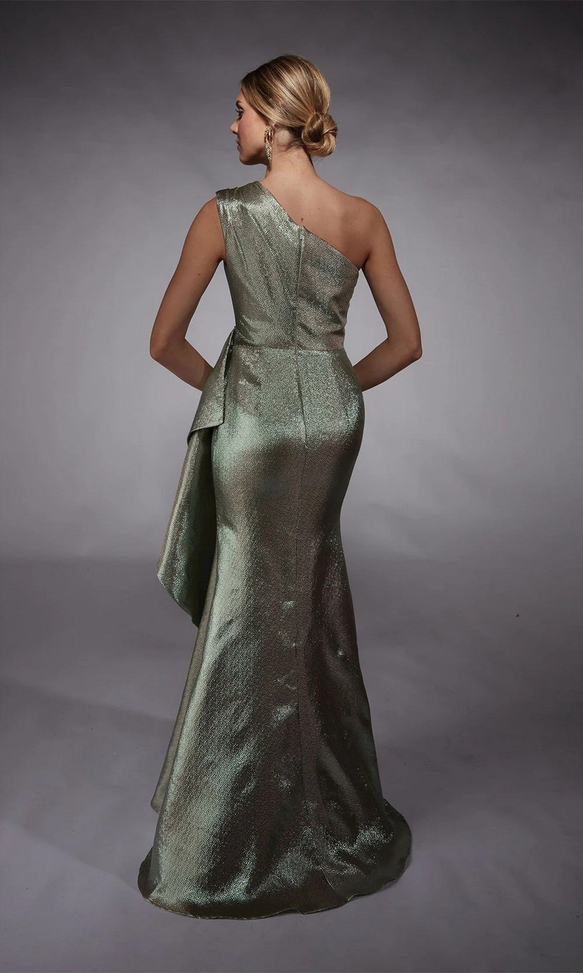 A woman stands on a gray backdrop wearing the Long Formal Dress: Alyce 27751—a metallic one-shoulder gown with an asymmetrical draped back, perfect as an elegant, modern mother-of-the-bride look.